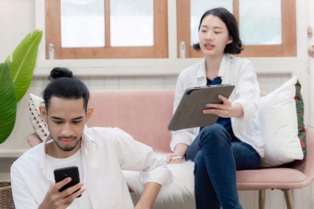 Two young individuals in casual medical attire are engaged in a study session. One person is sitting on a couch with a tablet in hand, while the other is seated nearby, focused on a smartphone. The relaxed environment suggests a collaborative study approach, common among medical students in the UK.