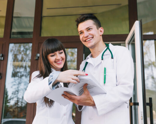 Two smiling medical students in white coats, standing outside a university building, reviewing study materials together. The male student wears a stethoscope, and both appear engaged in discussion, reflecting the collaborative and dynamic learning environment typical of studying medicine in the UK.