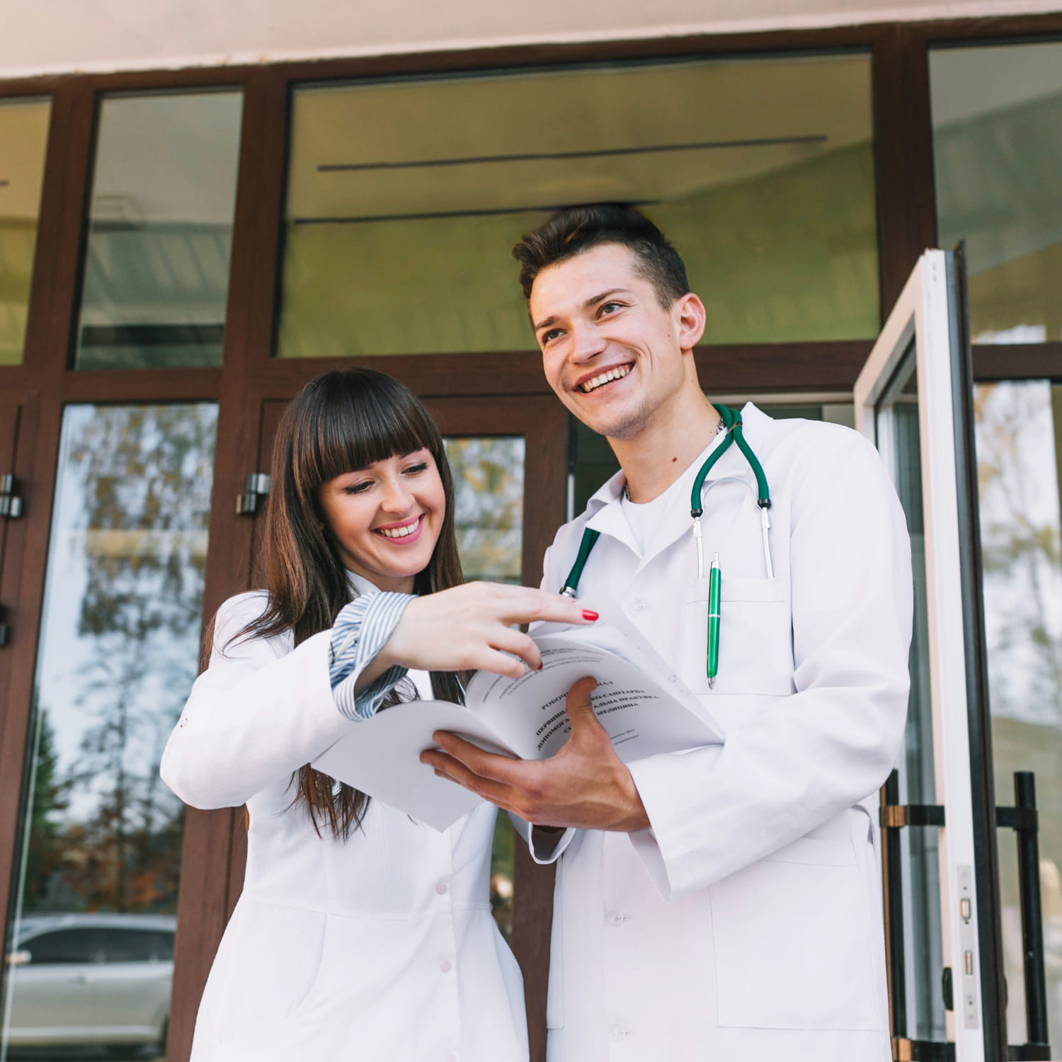 Two smiling medical students in white coats, standing outside a university building, reviewing study materials together. The male student wears a stethoscope, and both appear engaged in discussion, reflecting the collaborative and dynamic learning environment typical of studying medicine in the UK.