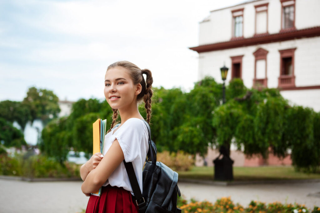 A student with braided hair, wearing a white top and red skirt, holds notebooks and stands outdoors in front of a building surrounded by greenery. The image depicts a young individual engaged in an affordable study abroad program in Ireland, with a backpack, ready for her academic journey.