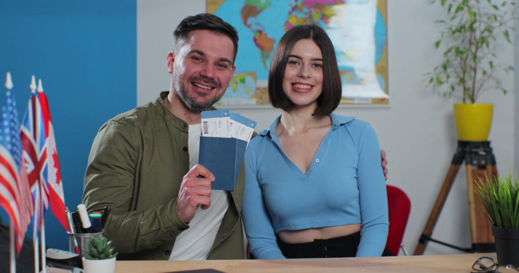 A smiling couple holding passports and flight tickets at an Expert PR Visa Consultancy office, surrounded by international flags and a world map in the background